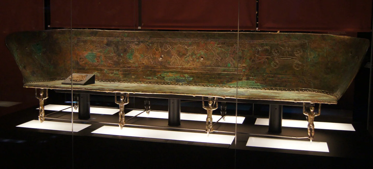 Lecho de bronce original de Hochdorf, V 86/3, en el Landesmuseum Württemberg; incisiones y soportes antropomorfos sobre ruedas visibles. Fotografía documental de 2016, no reconstrucción.