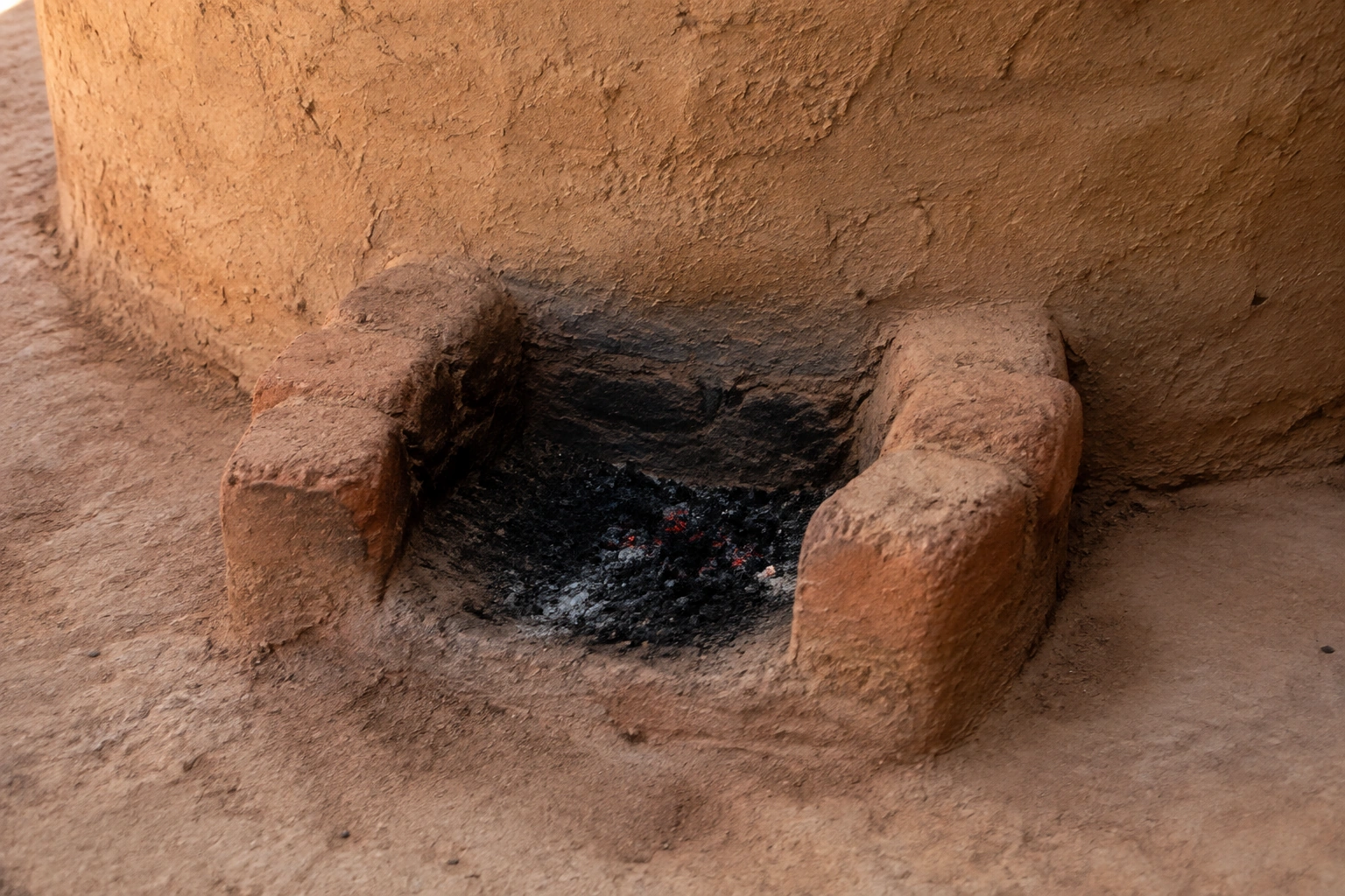 Hipótesis de detalle del hogar exterior de la casa SB de Jenne-jeno, Fase IV: dos muretes rubefactados junto a una pared curva; no reproduce un plano medido ni el alzado completo.