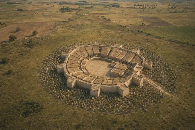 Reconstituição interpretativa aérea de Els Vilars I, cerca de 600 a. C.: recinto oval, casas retangulares adossadas e campo de pedras fincadas; alçados e coberturas hipotéticos.